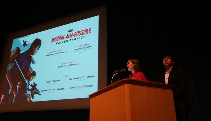 A man and woman presenting at a podium with a projector screen, discussing business innovation.