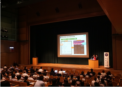 A man presenting business innovation to a large audience at a kaizen competition.