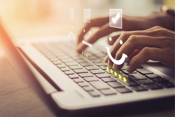 A person’s hands are shown typing on a laptop, with digital icons like stars, a checkmark, and a smiley face appearing above the keyboard, illustrating a high-quality customer experience facilitated by TMJP’s online BPO services.