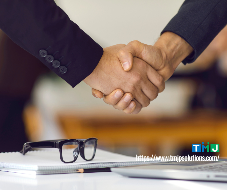 Two professionals in business suits shaking hands beside a desk with eyeglasses and a notebook, symbolizing Business English training driving stronger sales performance.