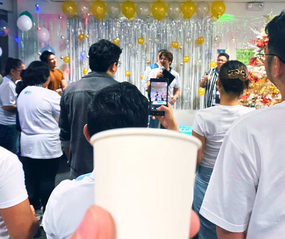 TMJP year-end party scene with people gathered in front of a silver and gold balloon backdrop, a decorated Christmas tree on the side, and someone holding a white paper cup in the foreground while another person records the event on a smartphone.